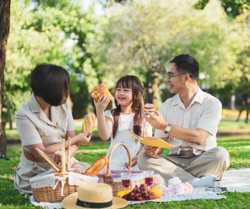 Happy Asian family enjoying a picnic together in a sunny park, sitting on a blanket with food and drinks, smiling and relaxed in the greenery