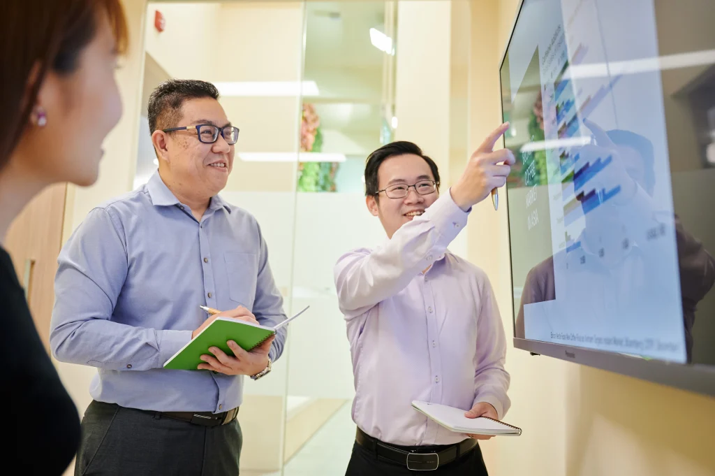 KH Roberts team members smiling and pointing at a large televised screen displaying data during a meeting.