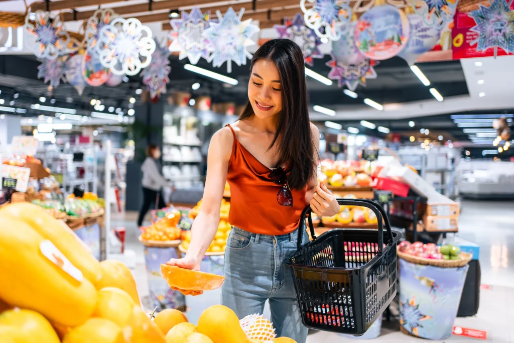 Young Asian Woman Holding Grocery Basket Choosing Fruits In Supermarket